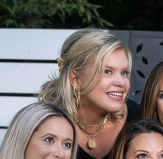 Group of women smiling outdoors with plants and a white wooden fence in the background.