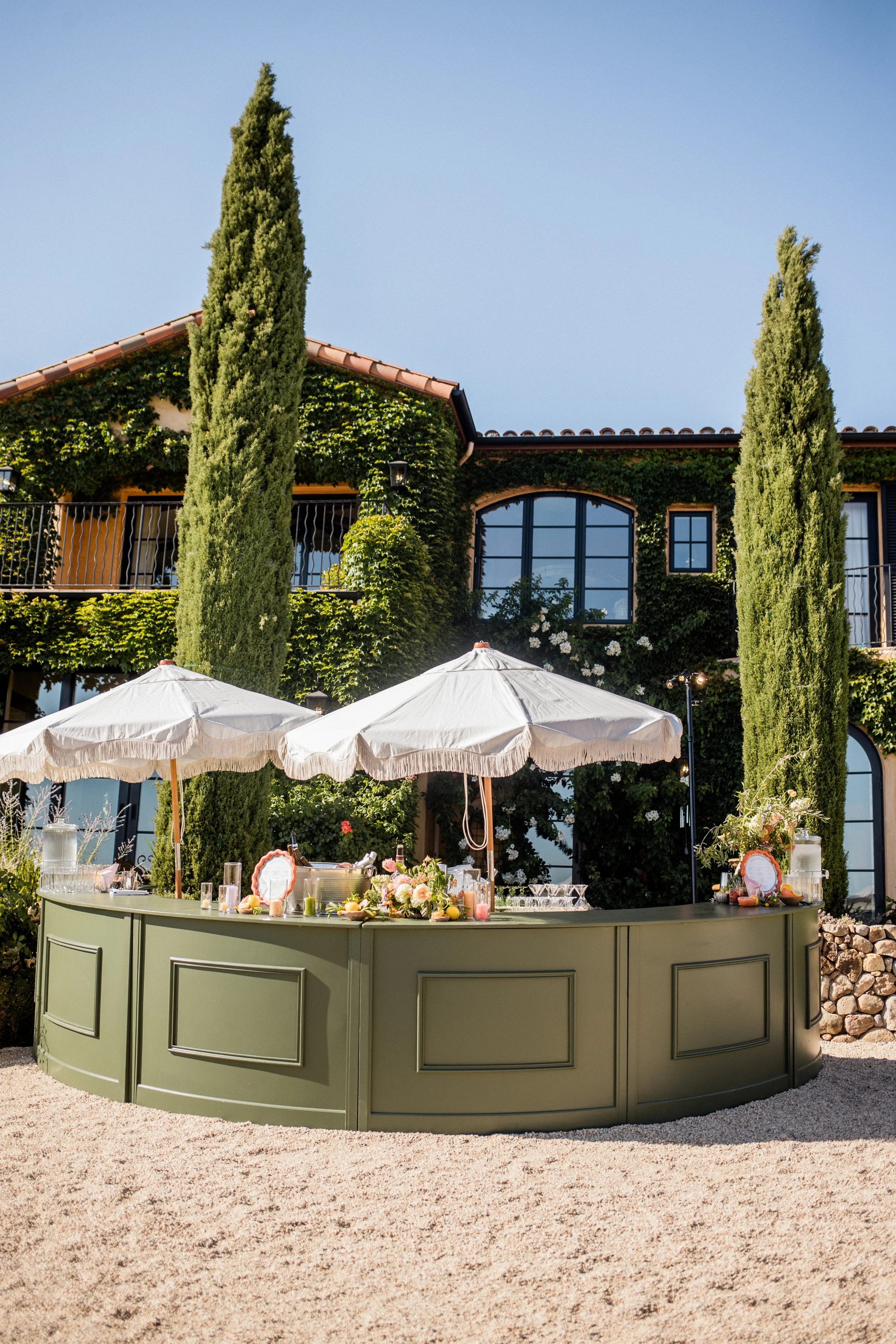 a bar for a wedding reception in sonoma with white umbrellas over it and an italian styled villa behind it