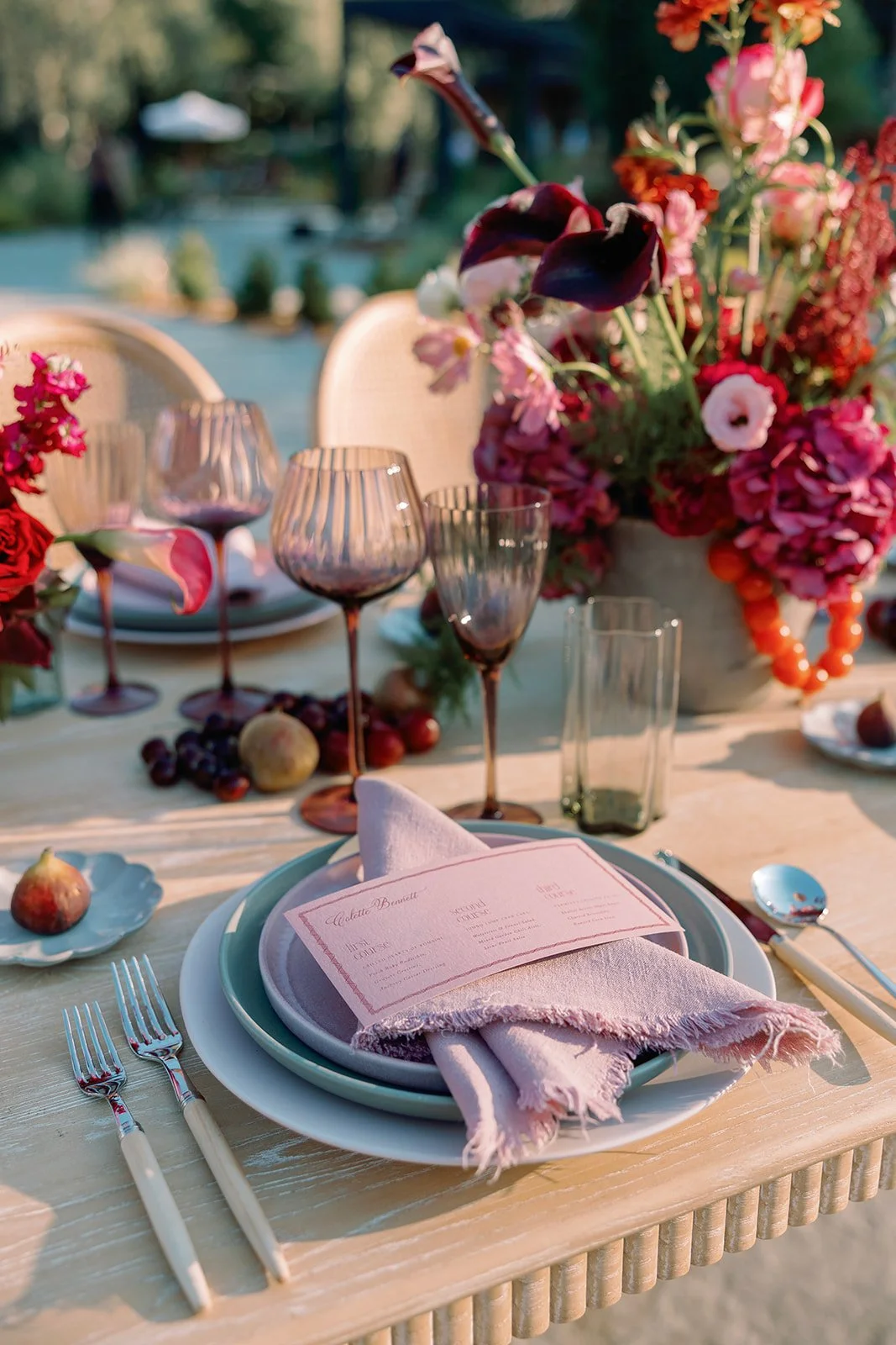 Elegant outdoor dining table set with pink and purple flowers, wine glasses, figs, grapes, and a menu, illuminated by warm sunlight.