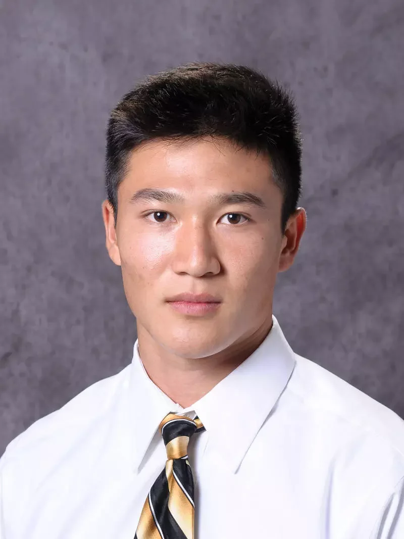 A young man with short dark hair wearing a white dress shirt and a striped black and yellow tie, looking directly at the camera with a neutral expression, against a gray textured background.