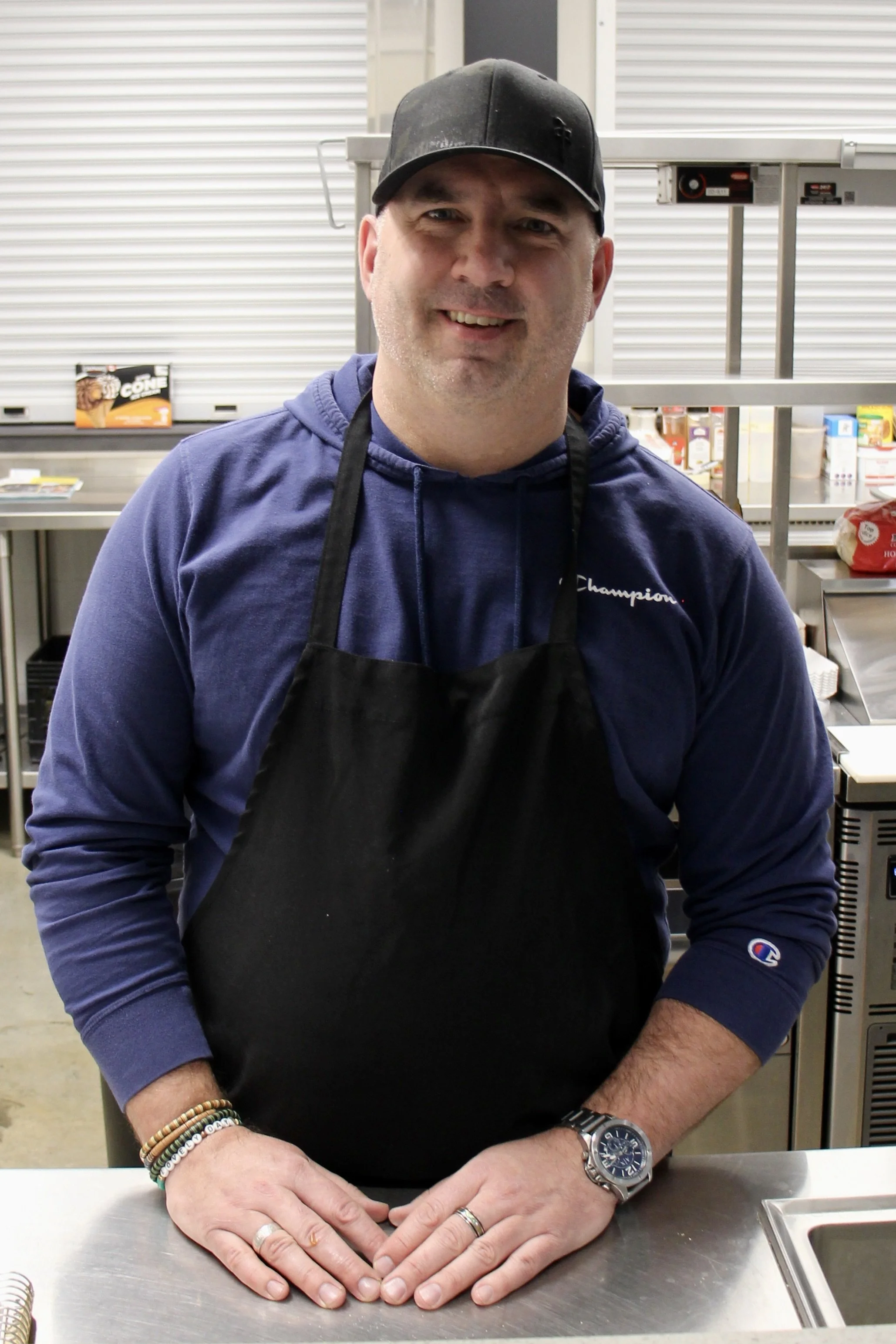 A man standing in a commercial kitchen, wearing a black apron, a blue hoodie, a watch, and a hat, smiling at the camera.