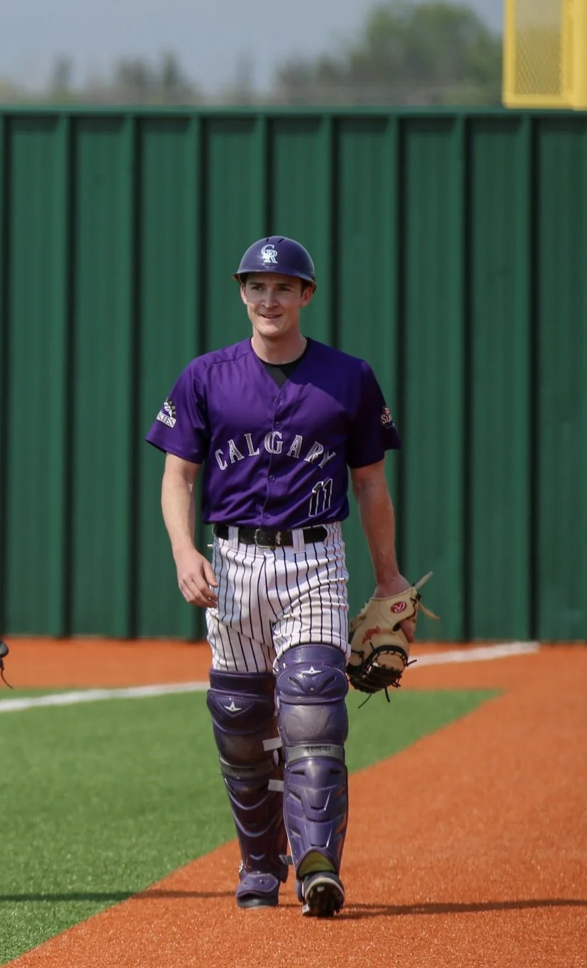 A young male baseball player wearing a purple Calgary jersey and striped pants, holding a baseball glove, walking on the field with a green outfield wall in the background.