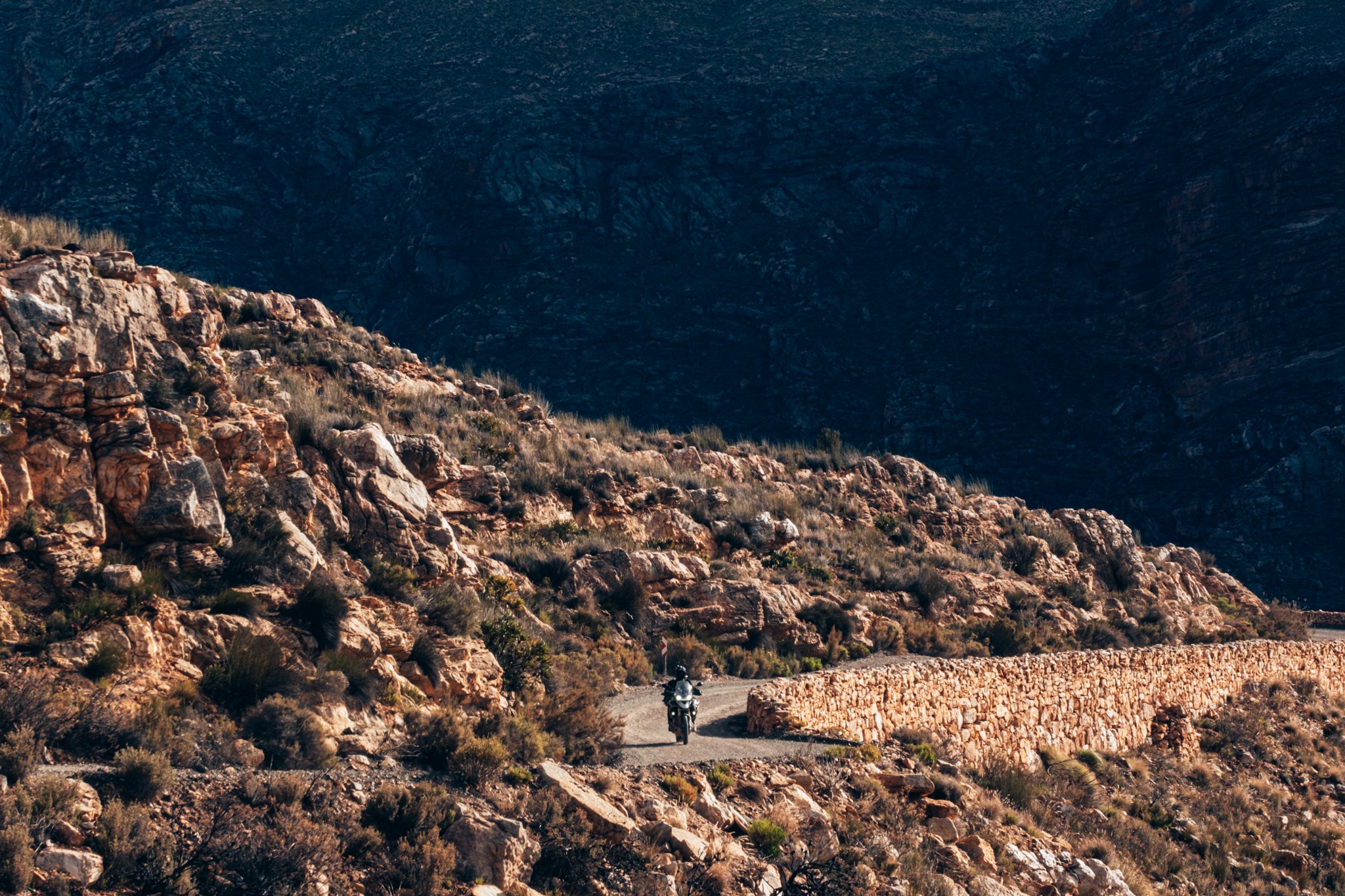 A motorcycle riding along a winding mountain road surrounded by rocky terrain and sparse desert vegetation.