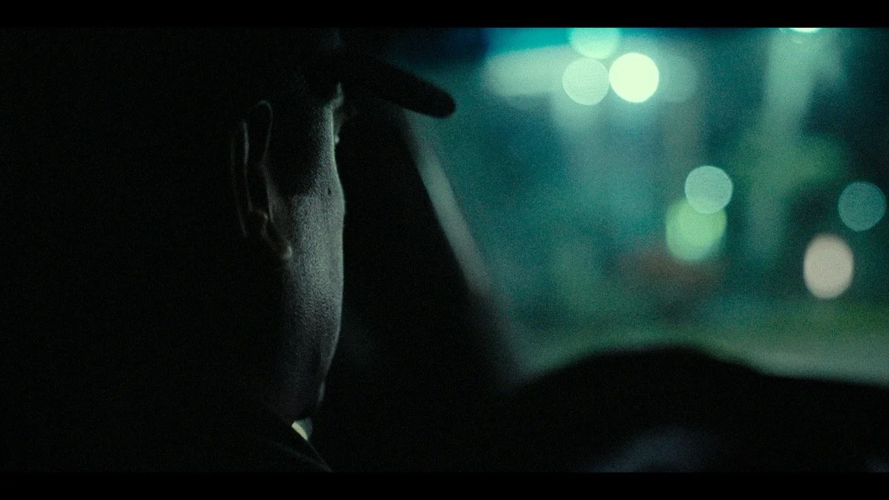 A person driving a car at night, viewed from the side with city lights blurred in the background.