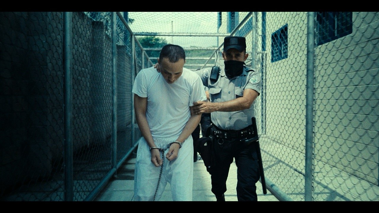 A detained man with hands chained and handcuffs, being escorted by a police officer through a fenced outdoor area.