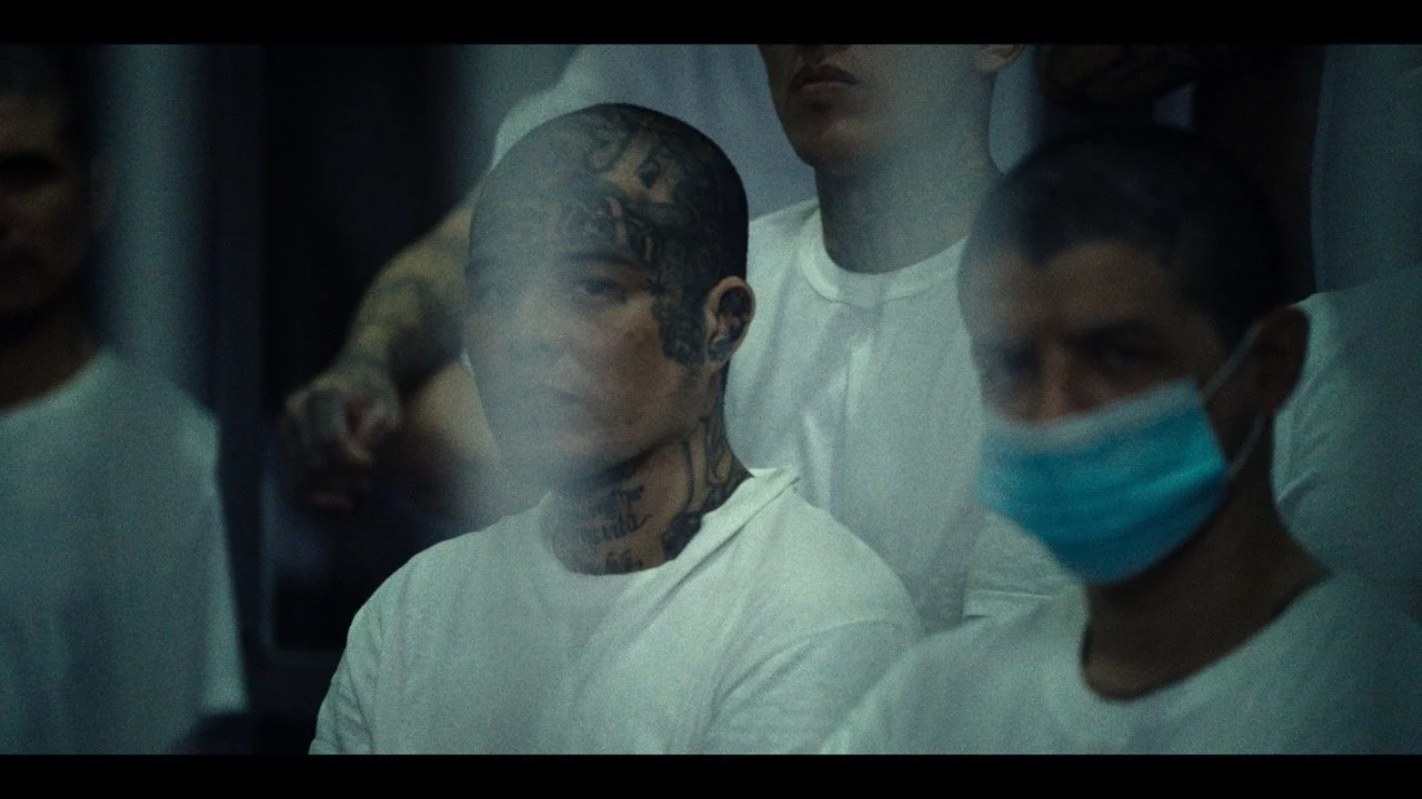 Group of people, including a tattooed person wearing a white shirt and a face mask, standing behind a glass window.