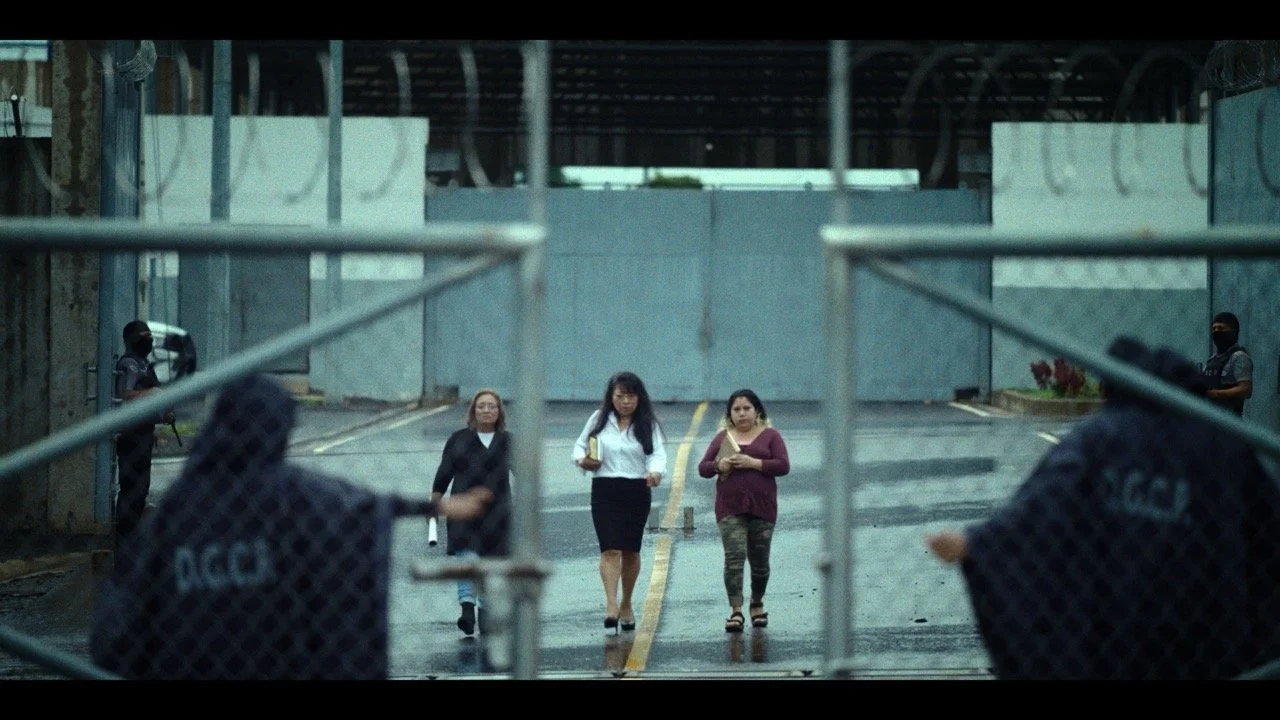 Three women walking on a street behind a metal barricade, with police officers on either side, in an urban area with a closed gate and building behind.
