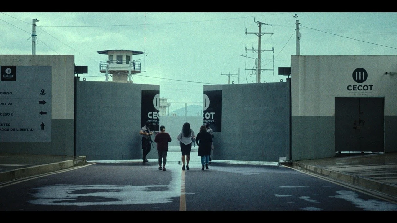 Group of four people walking toward CECOT gates at a facility entrance, with a watchtower and power lines in the background.