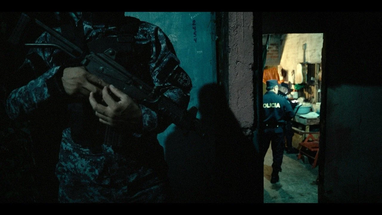 Military or police officers, one with a rifle, in a dark doorway, another in a hallway leading to a room with a person wearing a police uniform, in an indoor setting.