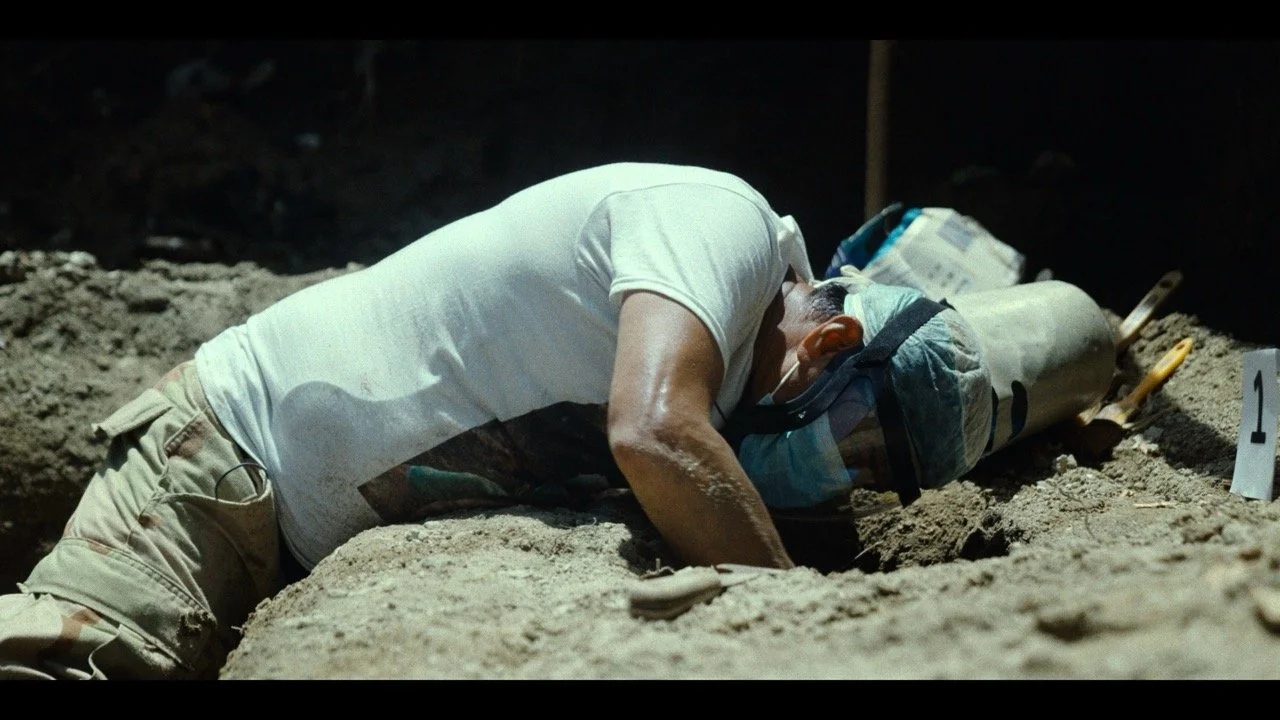 A person is lying face down on the ground at an excavation site, with their head resting on a cylindrical object, possibly a archaeological artifact, and wearing a hair cover and gloves. A numbered marker is nearby.