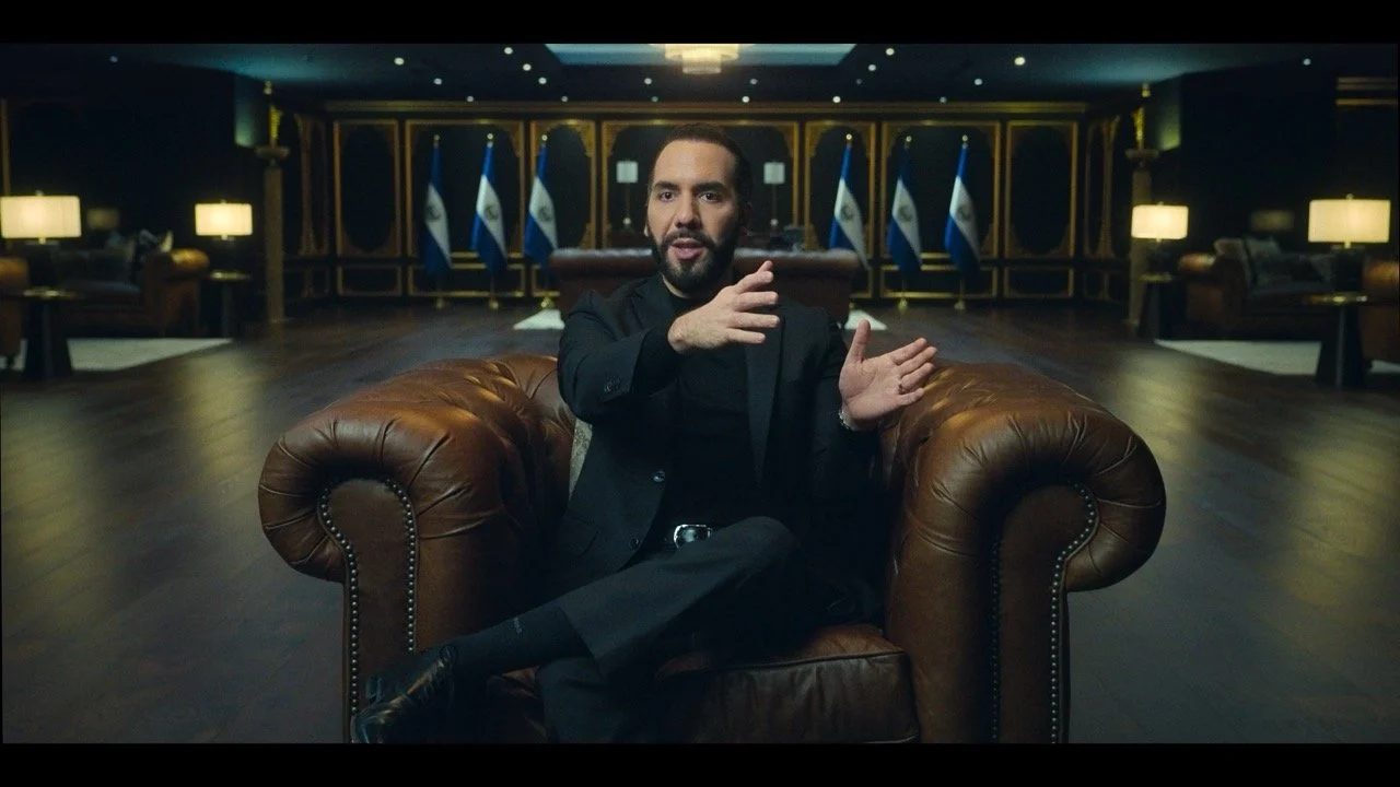 A man with a beard and dark hair, dressed in a black suit, sitting on a brown leather armchair in a dimly lit room with a dark wooden floor and walls, with multiple flags in the background.