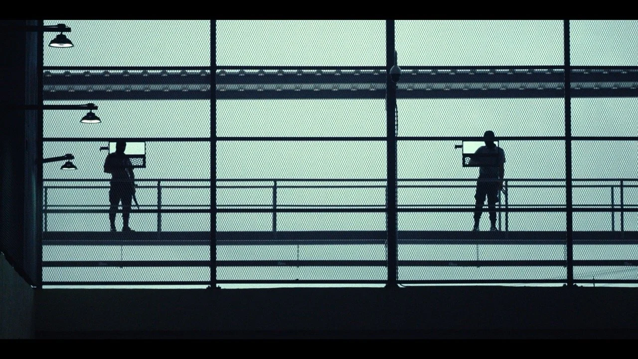 Two workers on a high, enclosed outdoor balcony with mesh fencing, each working on equipment at their stations.