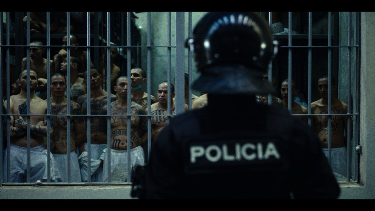 A police officer wearing a helmet and a jacket with 'POLICIA' on the back is standing in front of a jail cell with multiple shirtless men with tattoos inside.