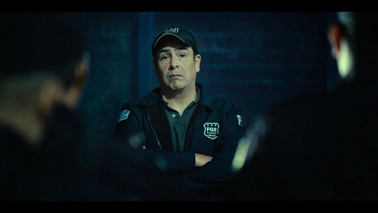 A police officer with crossed arms, wearing a dark uniform and cap, stands in a dimly lit setting, speaking to a group of people.