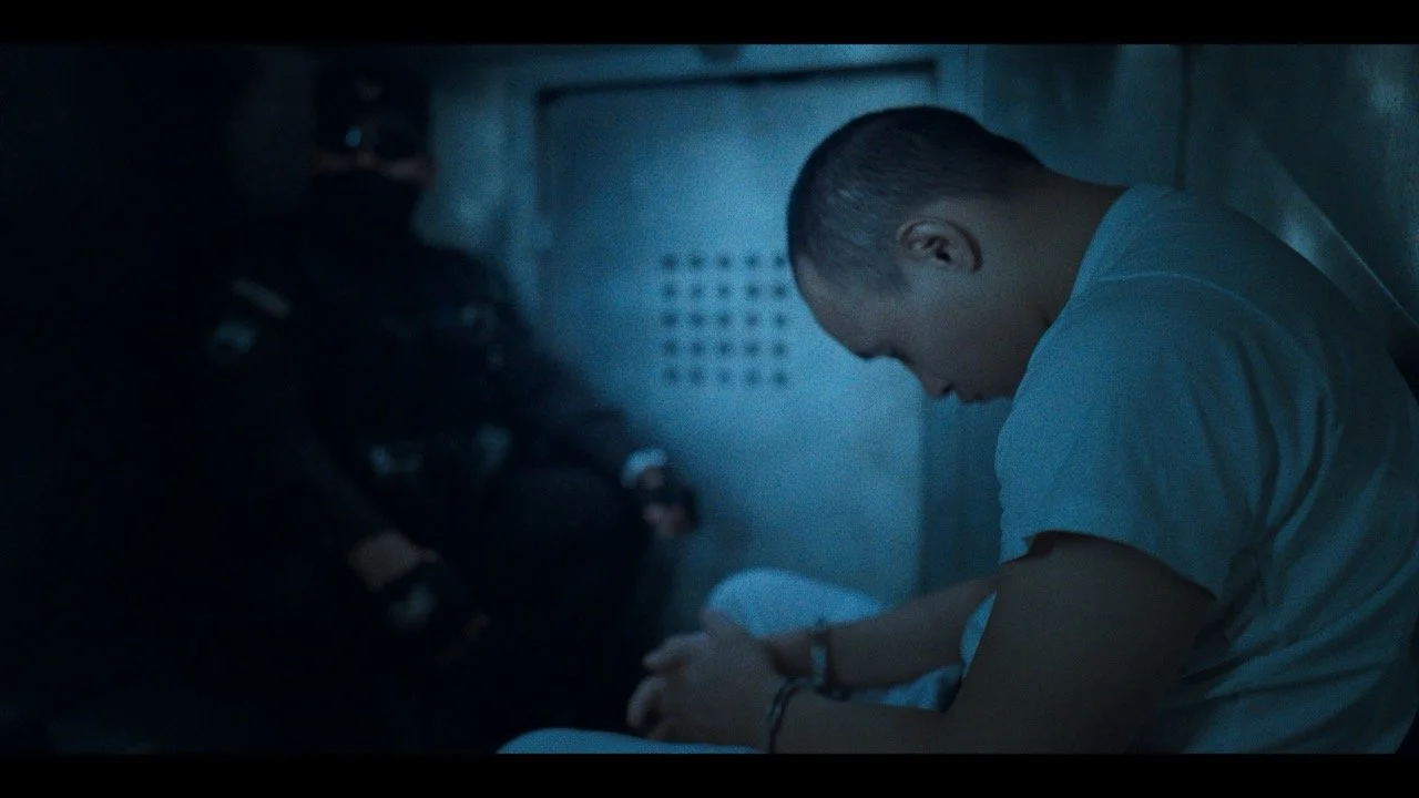 A young man with a shaved head sitting with his head bowed in a gray T-shirt, in a dimly lit room, with two police officers sitting in the background.