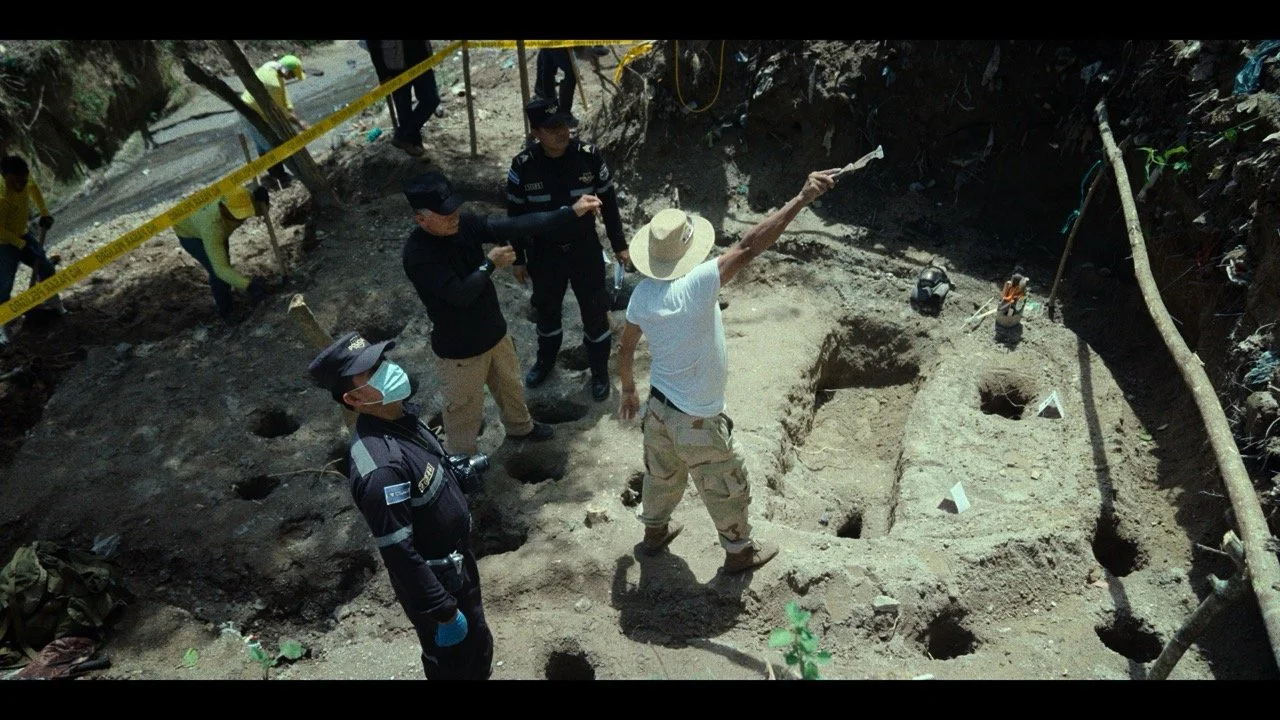 Archaeologists excavating an archaeological site with various holes in the ground, while police and investigators observe and discuss the site.