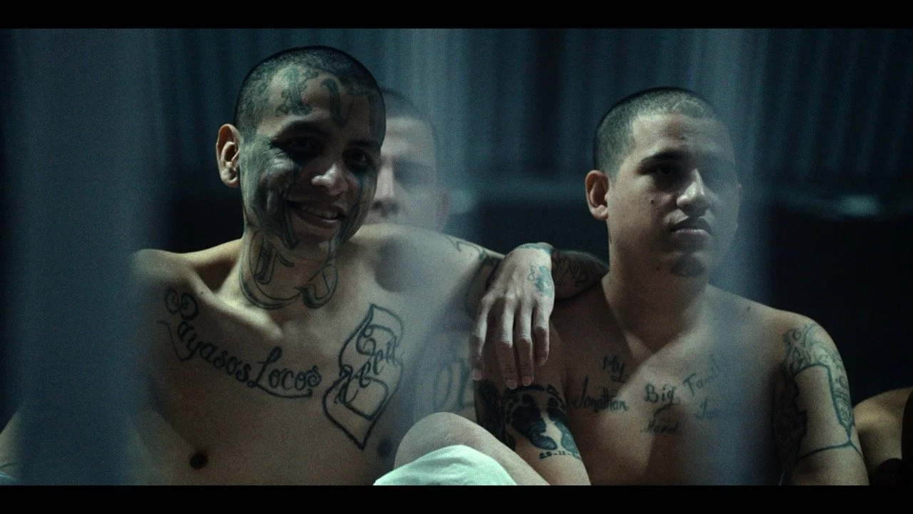 Two shirtless men with tattoos sitting next to each other behind a glass window, with one smiling and the other looking serious, in a dimly lit room.