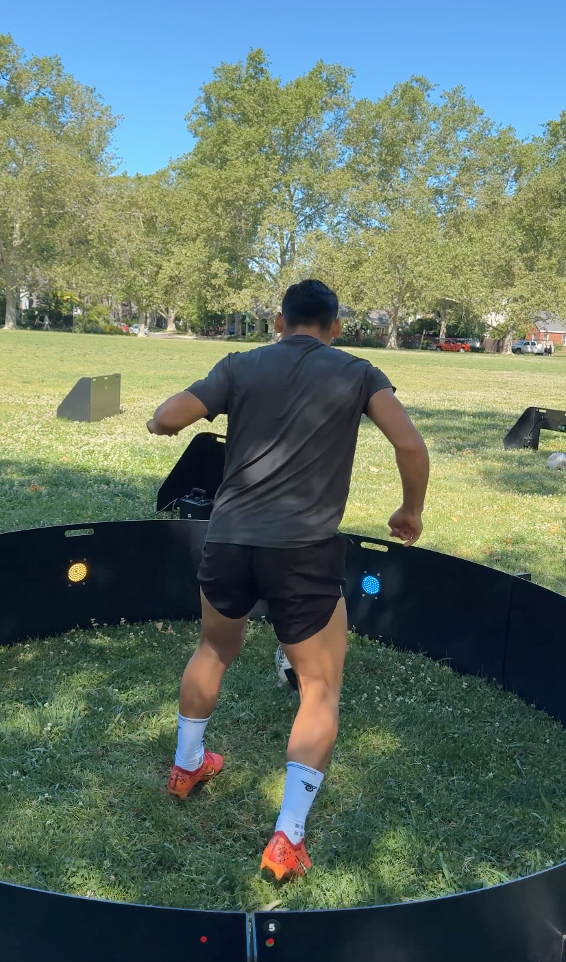 A man is playing a sport called footgolf outdoors in a grassy park with trees and blue sky in the background.