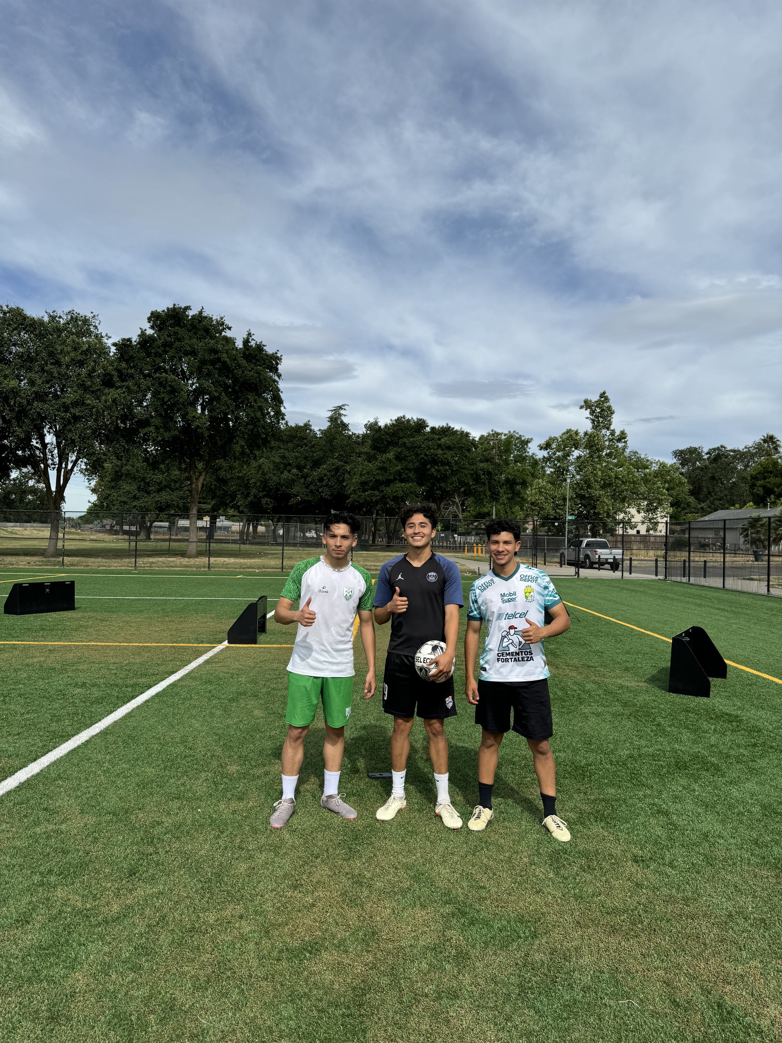 Three young men standing on a soccer field, smiling and giving thumbs-up signs, with trees and a cloudy sky in the background.