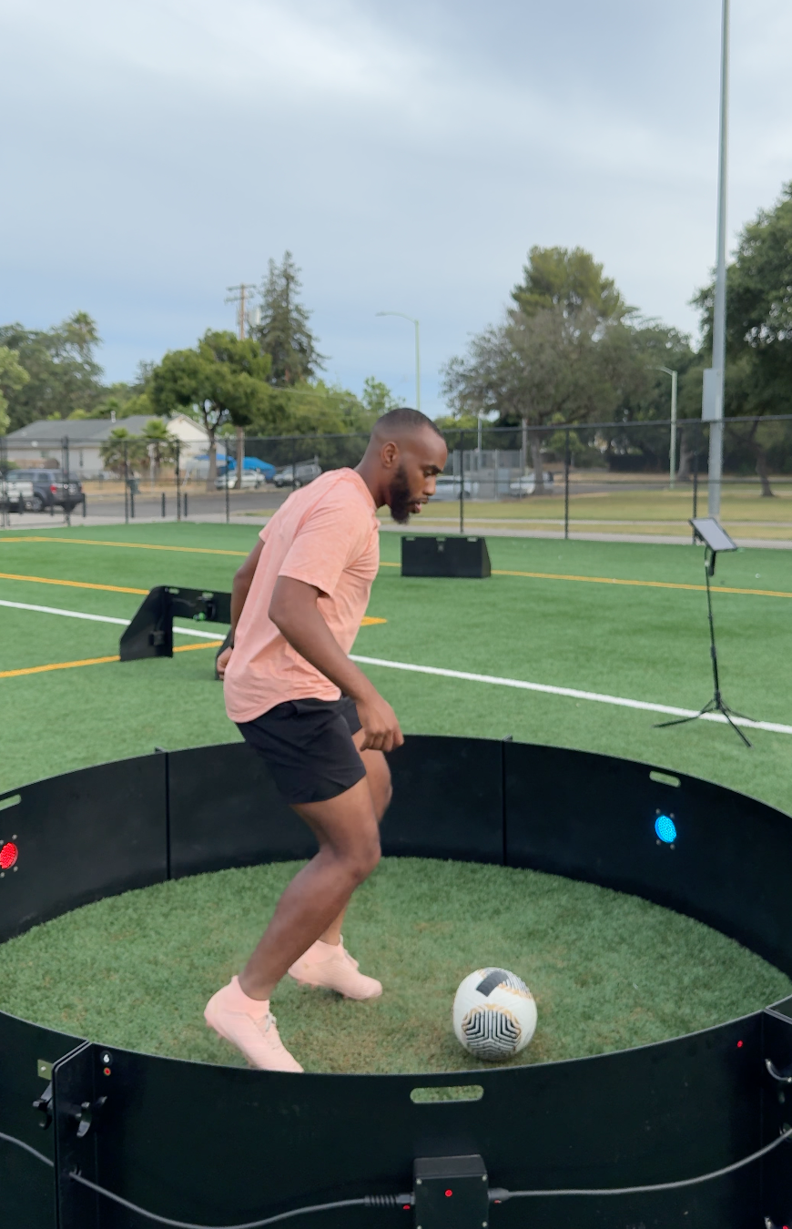 A man practicing soccer skills inside a large interactive training circle on a grassy field, with a soccer ball at his feet and digital sensors on the perimeter.