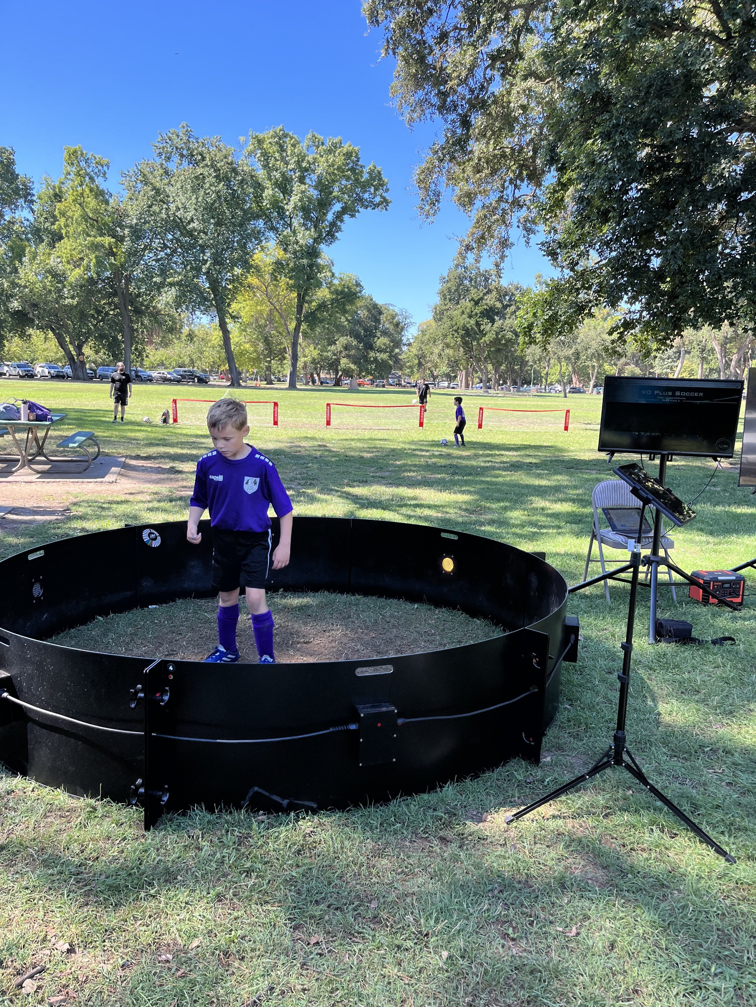 A young boy standing inside a black, circular mechanical or electronic device outdoors in a park on a sunny day. In the background, other children are playing soccer on a grassy field, surrounded by trees.