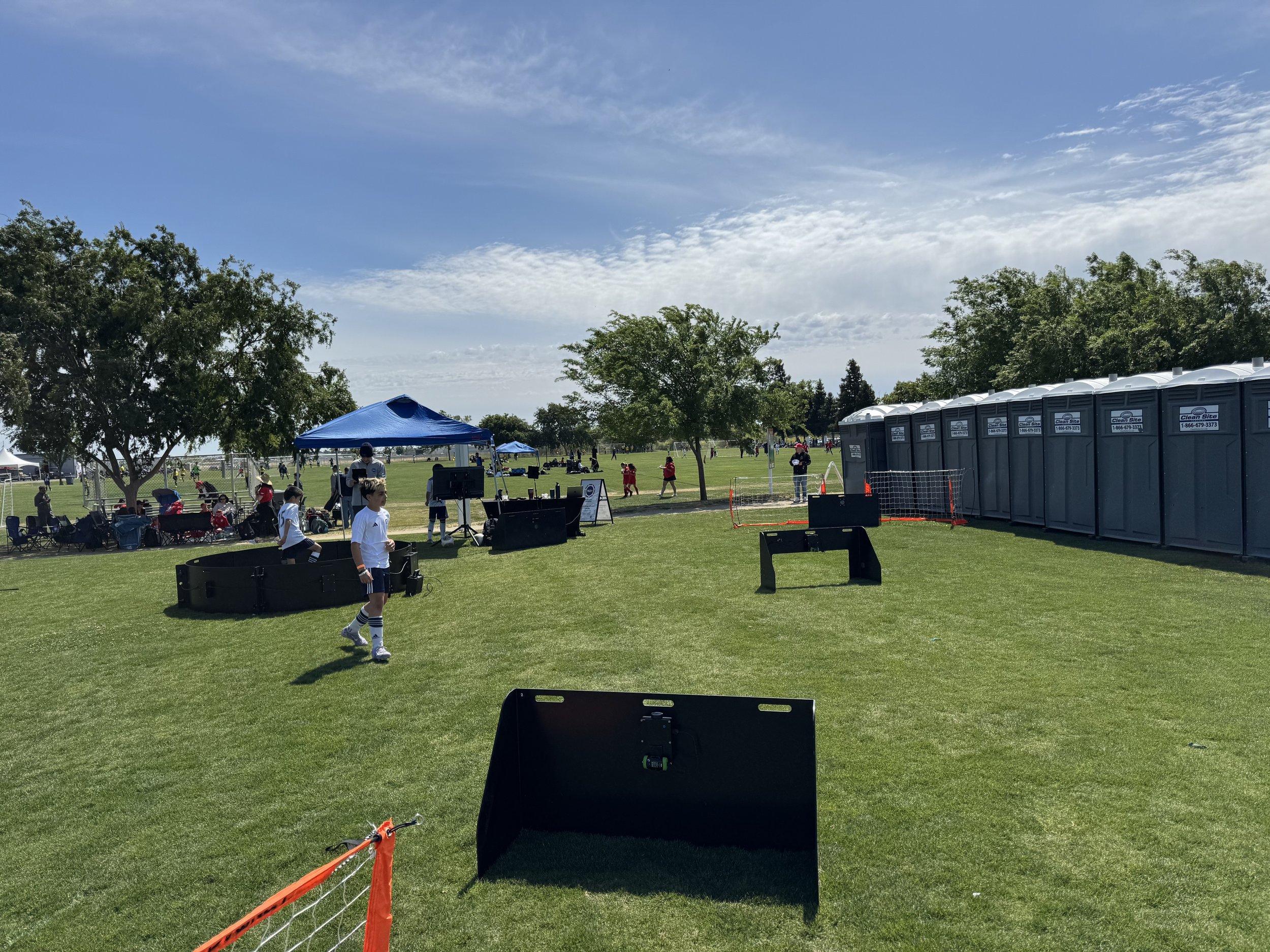 Outdoor park with portable toilets, children playing, and a shaded tent area on a sunny day.