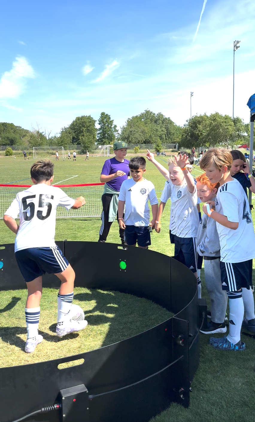 Children playing soccer outdoors on a sunny day, with some children gathered around a robot allowing a game of footsoccer.
