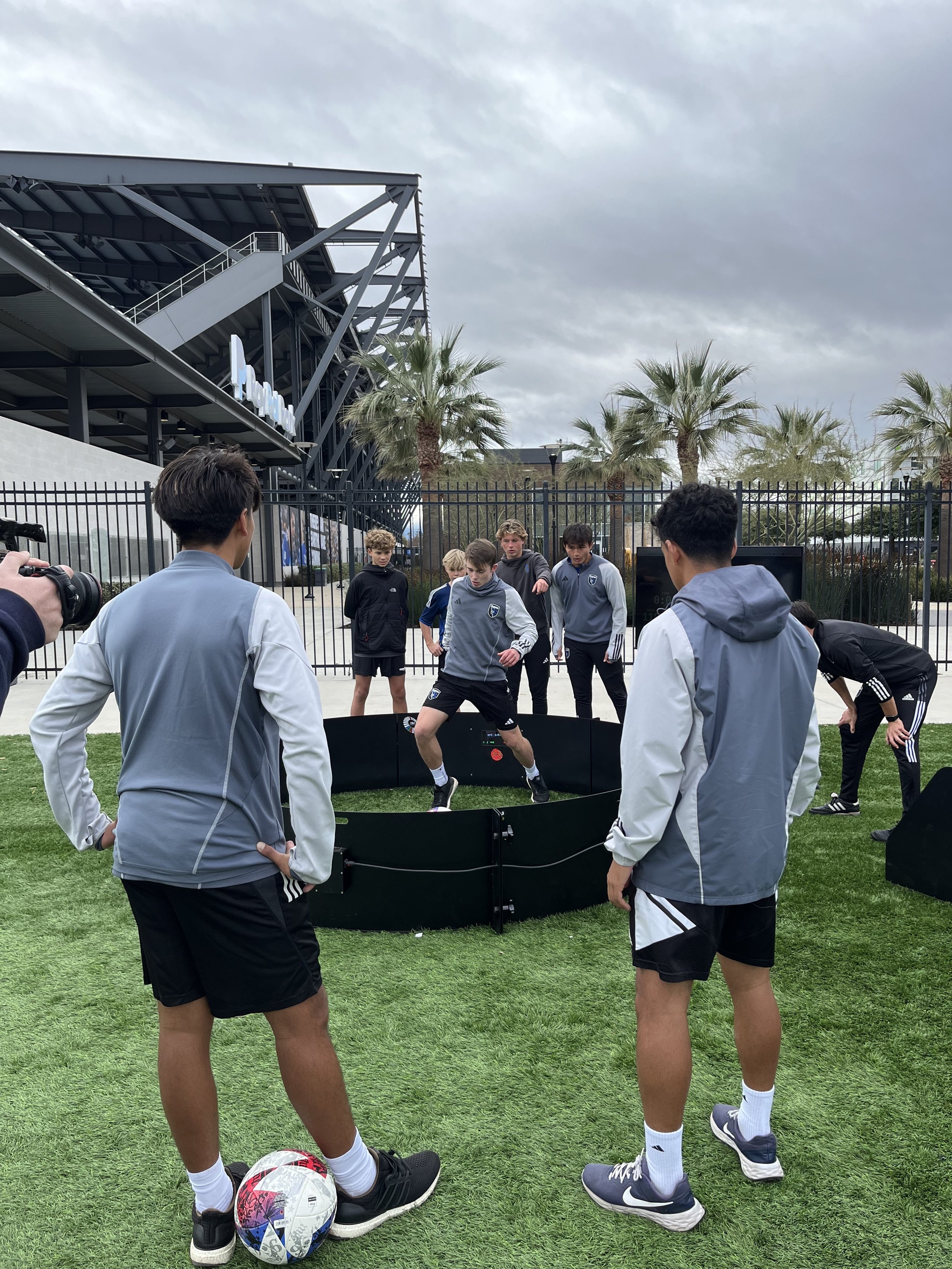 Group of young soccer players practicing dribbling drills on a field with a coach, with a stadium and palm trees in the background under cloudy skies.