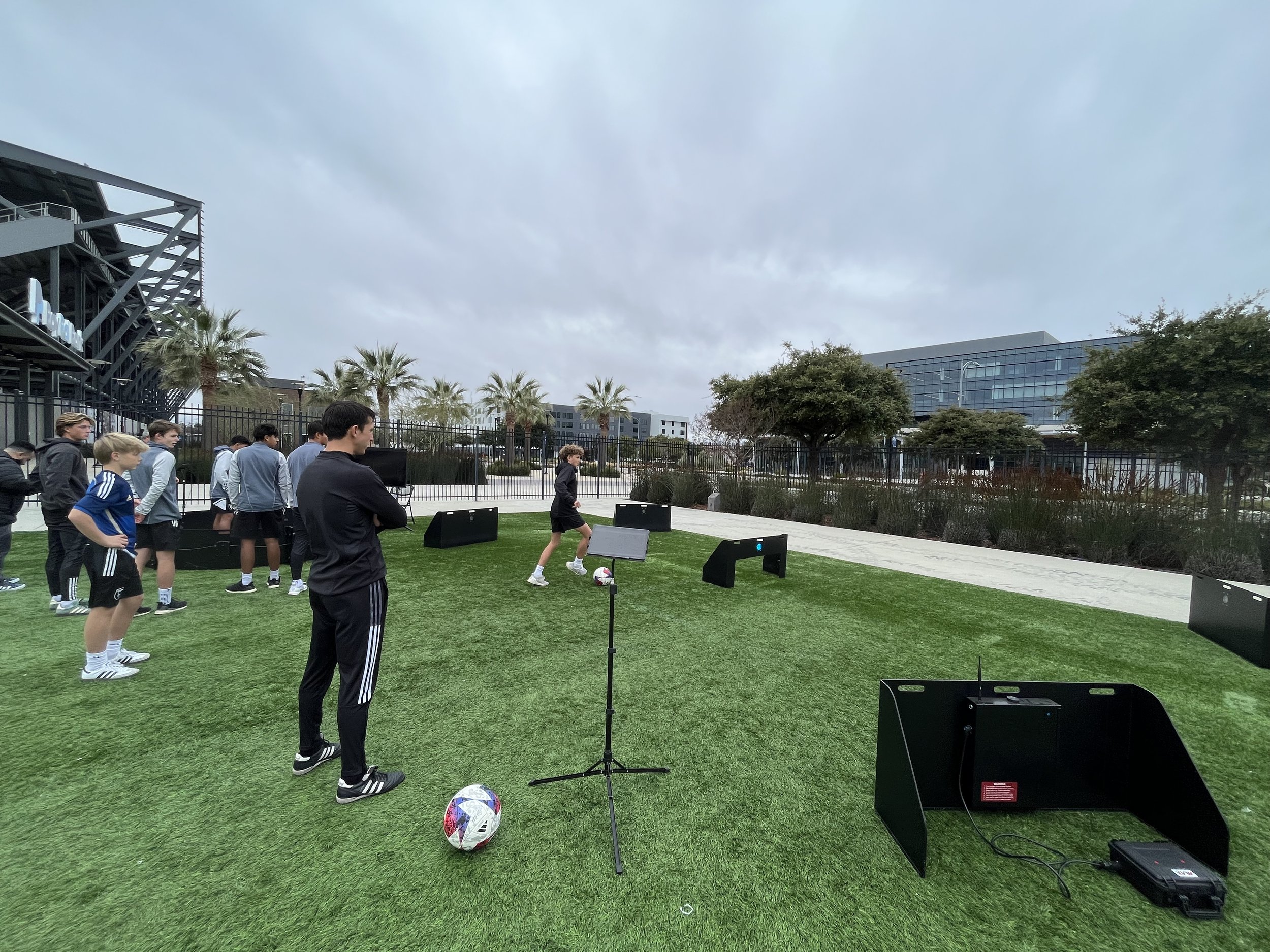 Group of children and coaches practicing soccer on a green field outdoors with palm trees and modern buildings in the background, overcast sky.