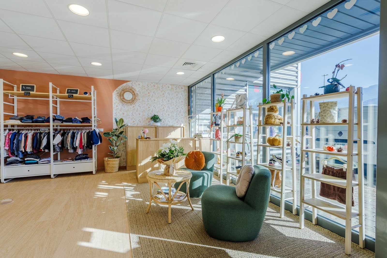 Boutique avec des étagères en bois blanc décorées avec des plantes en pot, des coussins et des décorations, un coin salon avec deux fauteuils verts, une table ronde avec un vase de fleurs, et un mur orange avec des étagères de vêtements pour enfants.