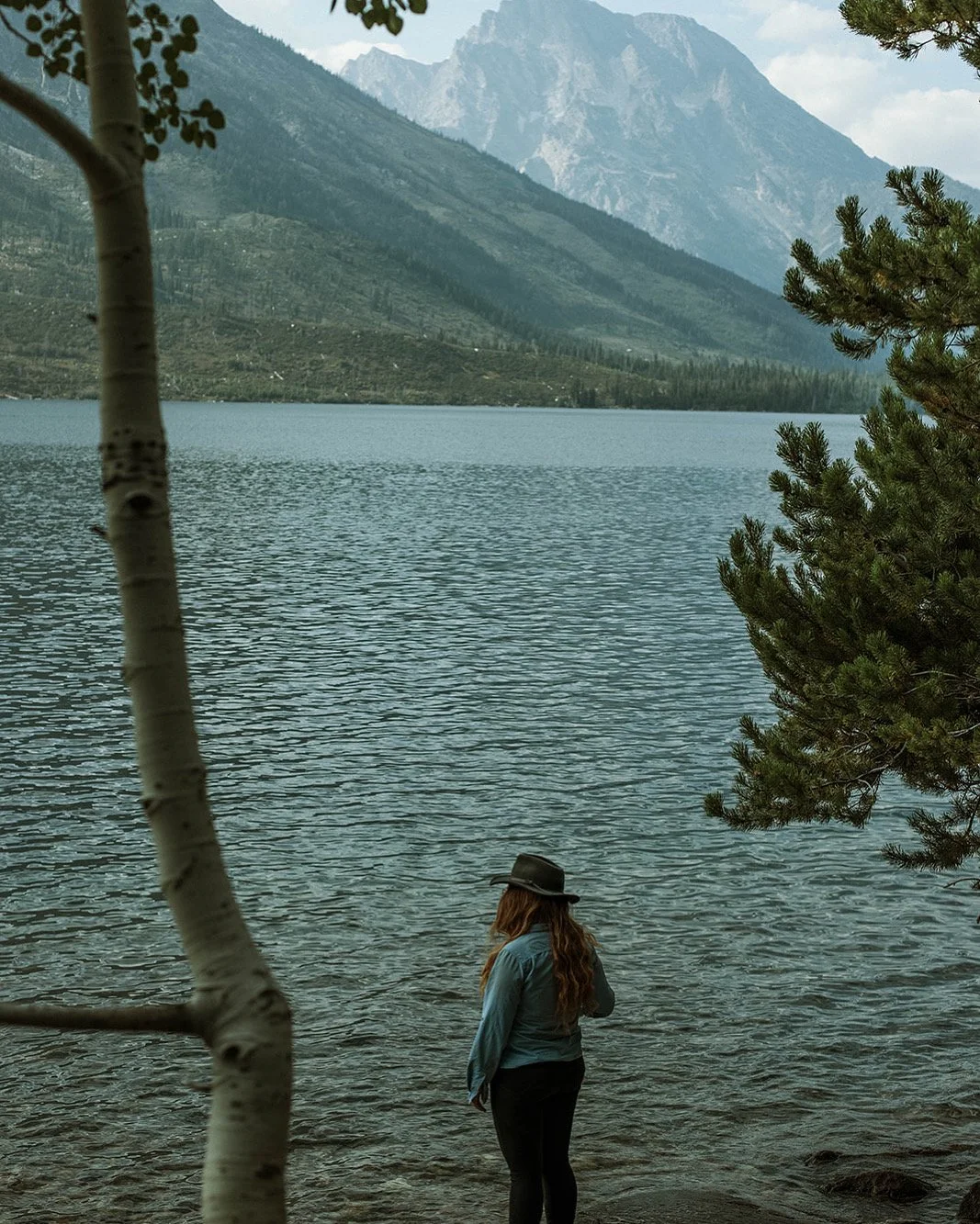 A woman with long hair, wearing a hat and a gray jacket, standing near the edge of a lake with mountains in the background, surrounded by trees.