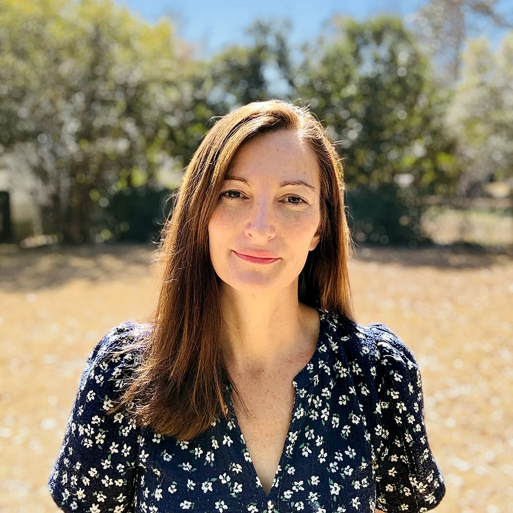 A woman with long brown hair smiling outdoors on a sunny day, with trees and a blue sky in the background.