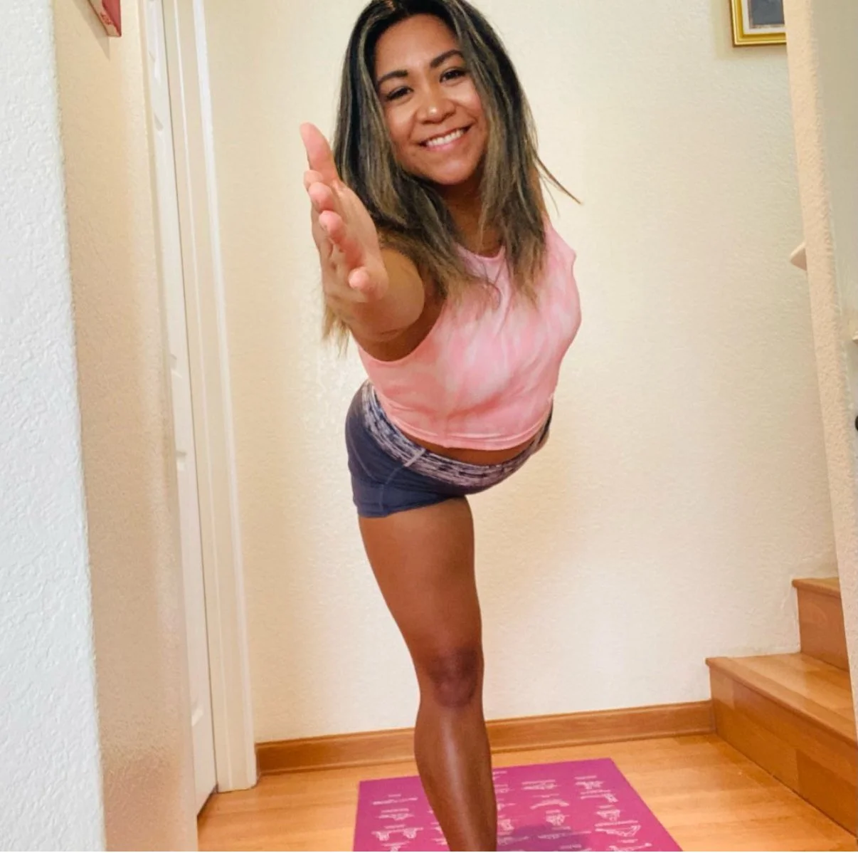 Woman doing a yoga pose on a pink mat indoors, dressed in a pink tie-dye shirt and dark shorts, smiling at the camera.