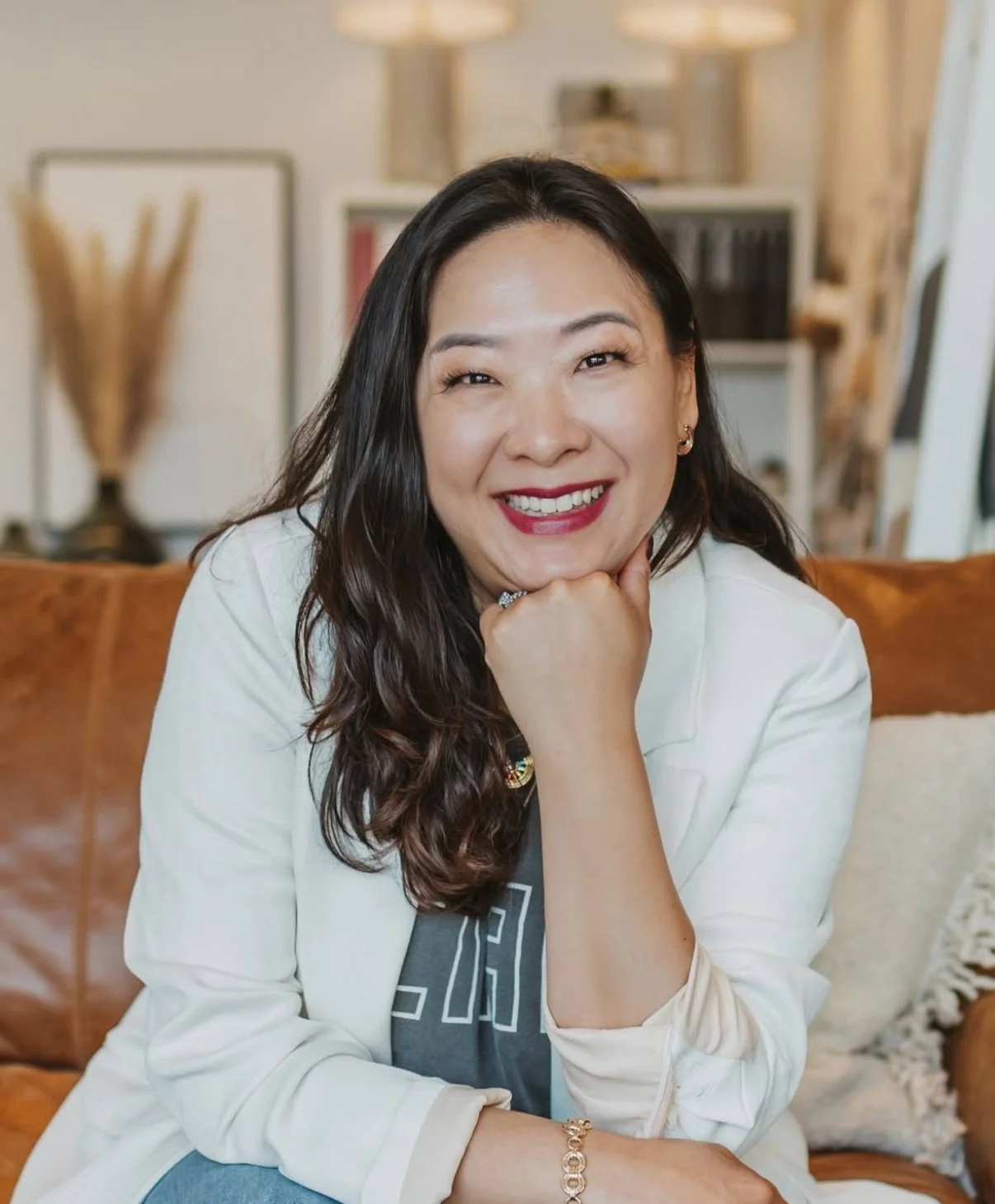 Smiling woman with long dark hair wearing a white blazer, sitting on a brown leather sofa in a cozy living room.