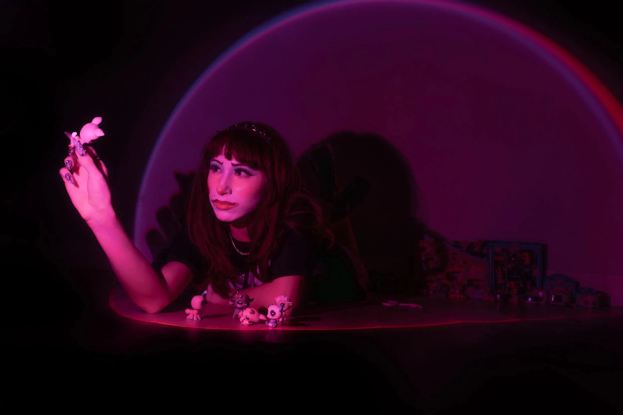 A woman with red hair and bold makeup lying on the floor in a dark room illuminated by pink and purple neon lights, surrounded by small toy figures.