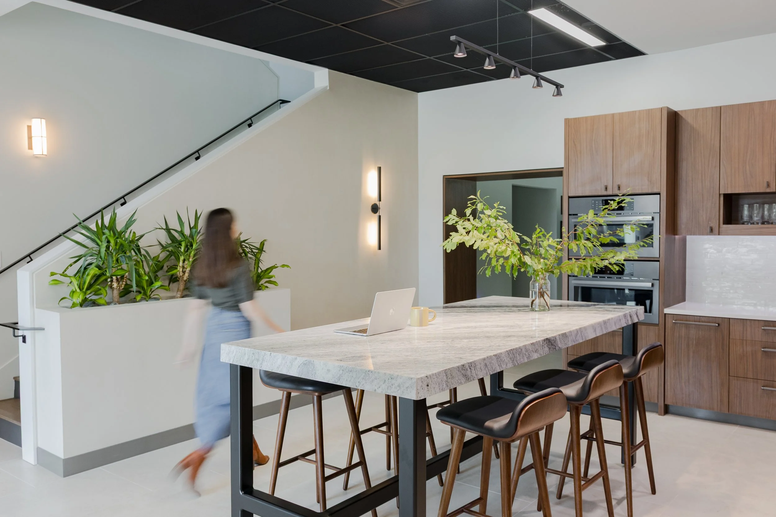 Modern kitchen with a large marble island, wooden cabinets, plants, and a person walking past.