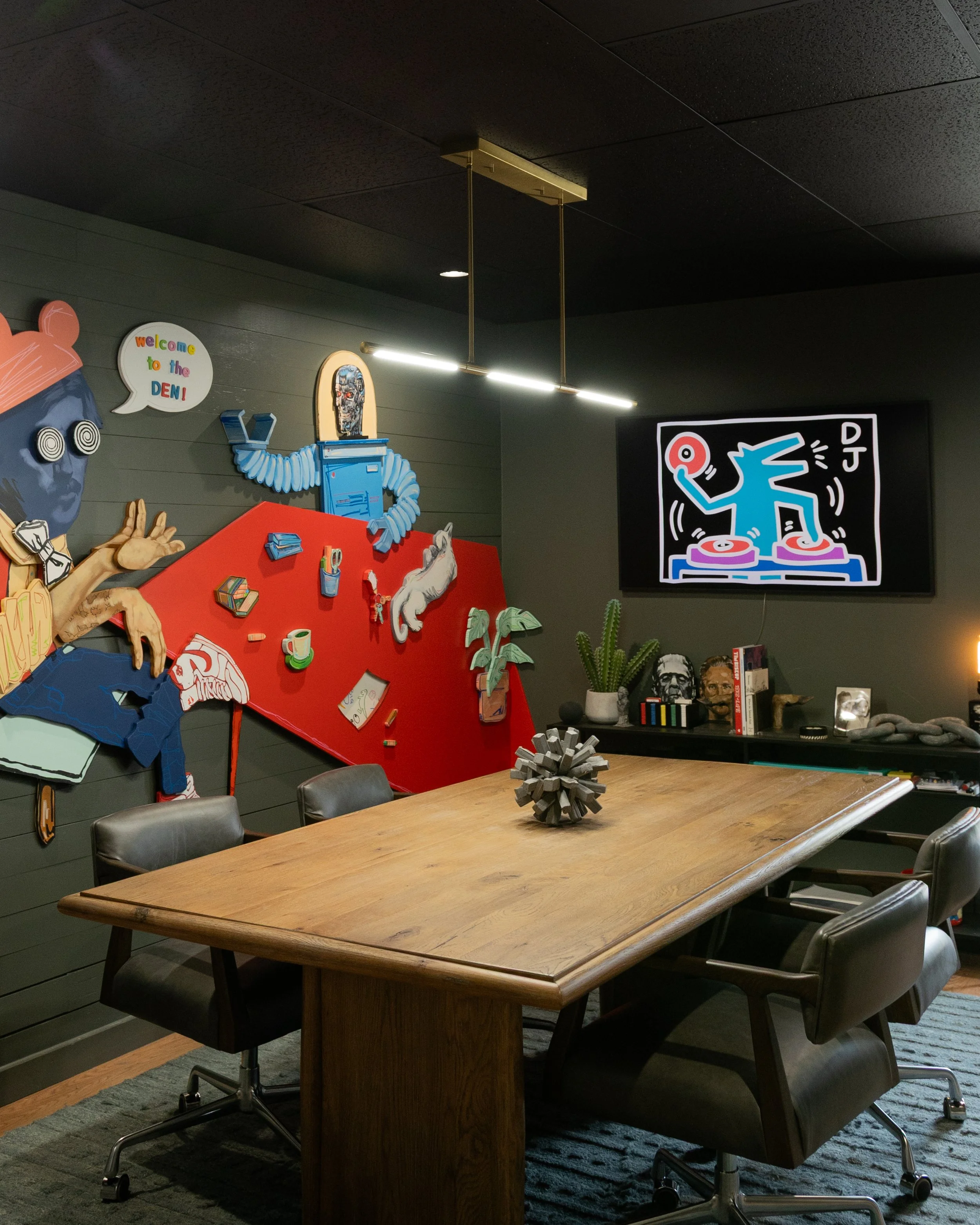 A conference room with a wooden table surrounded by six chairs. The room has dark paneled walls, a black ceiling, and various decorations including a red bulletin board with items pinned on it, plants, and wall art. Overhead lighting illuminates the 