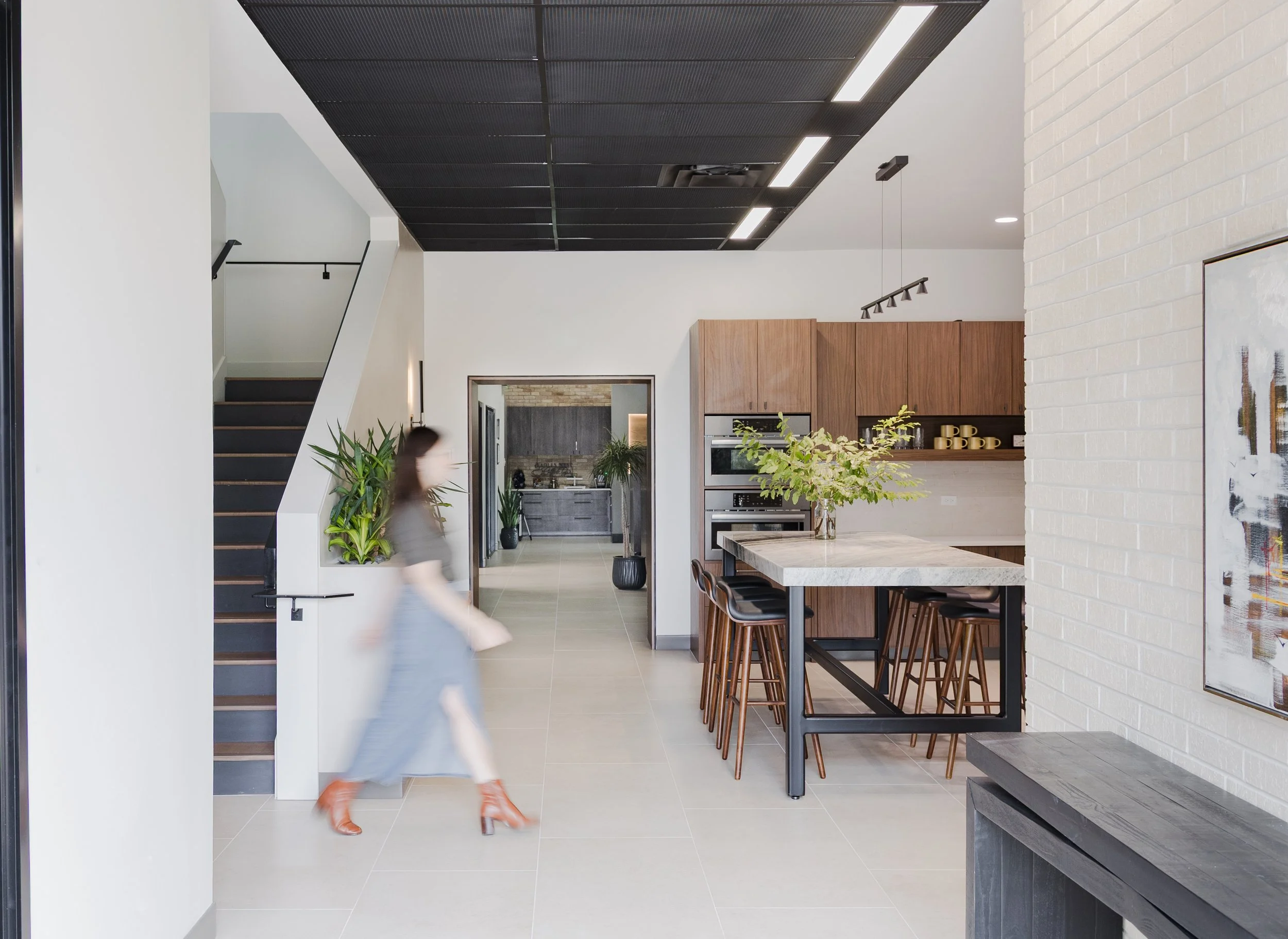 Modern kitchen and dining area with a white brick wall, wooden cabinets, and a large island with bar stools. An open doorway leads to another room with gray cabinetry and green plants. A person is walking past, appearing blurred.