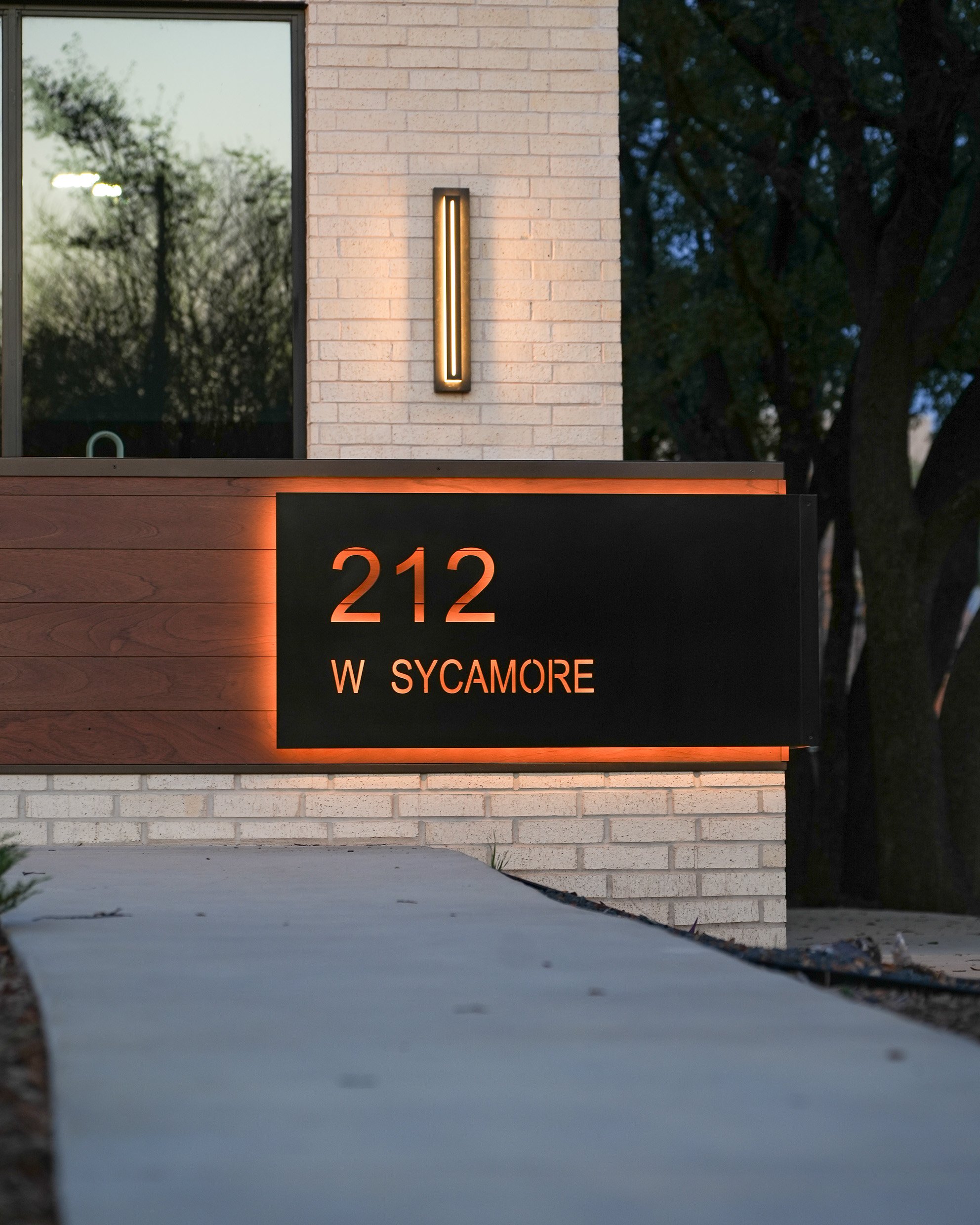 Close-up of a modern exterior wall and signage with street address 212 and W Sycamore, a lit vertical wall sconce fixture, on a brick building at dusk with trees in the background.