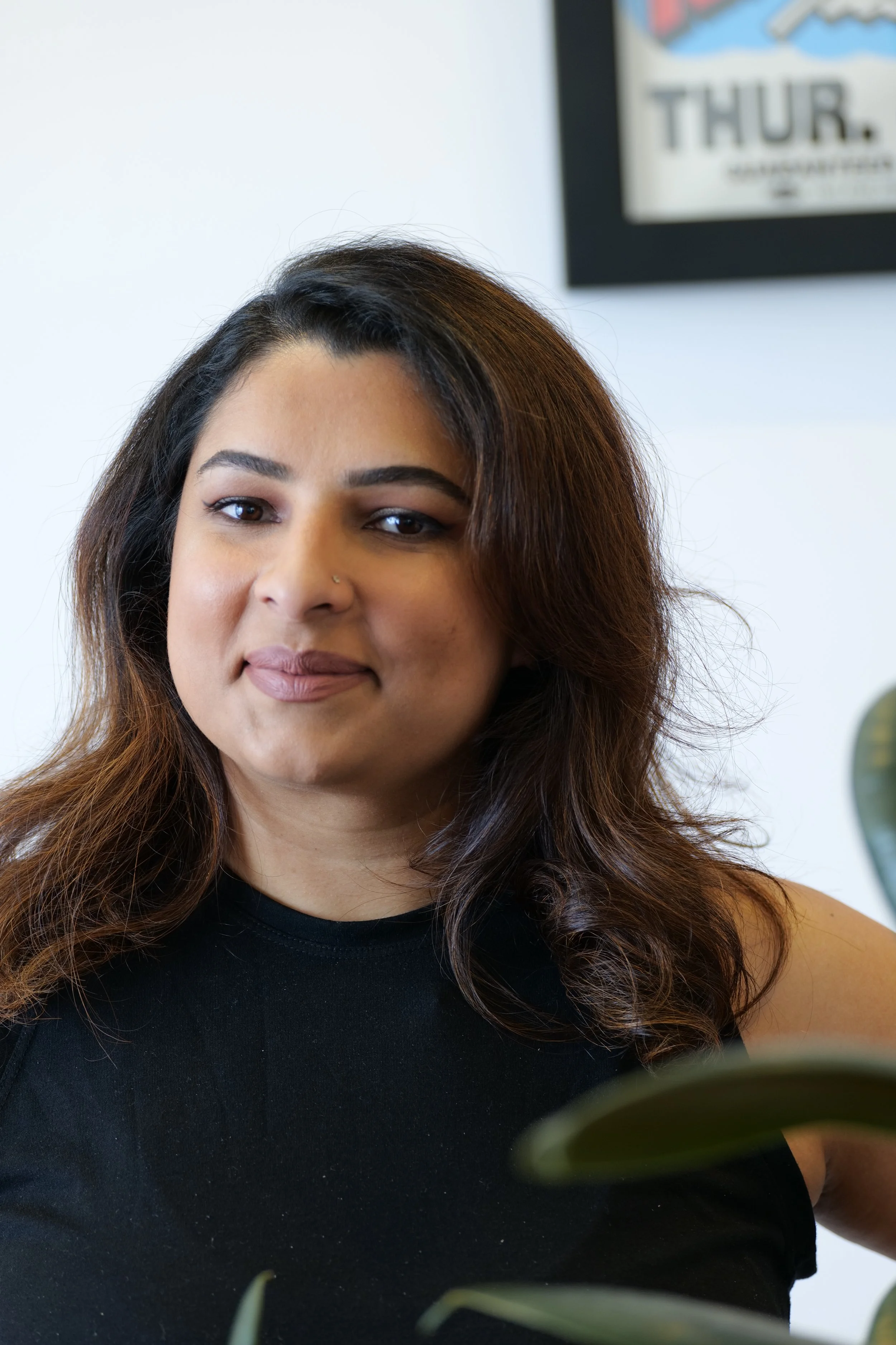 A woman with wavy brown hair and a nose piercing, wearing a black sleeveless top, smiling subtly indoors, with a framed poster in the background.