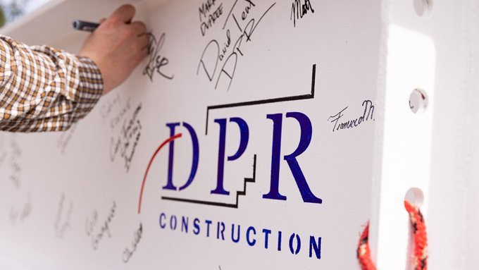 Team members sign a topping out beam around a stenciled DPR Construction logo