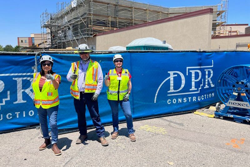 Three people in PPE standing in front of a blue fence wrap with DPR Construction logo.