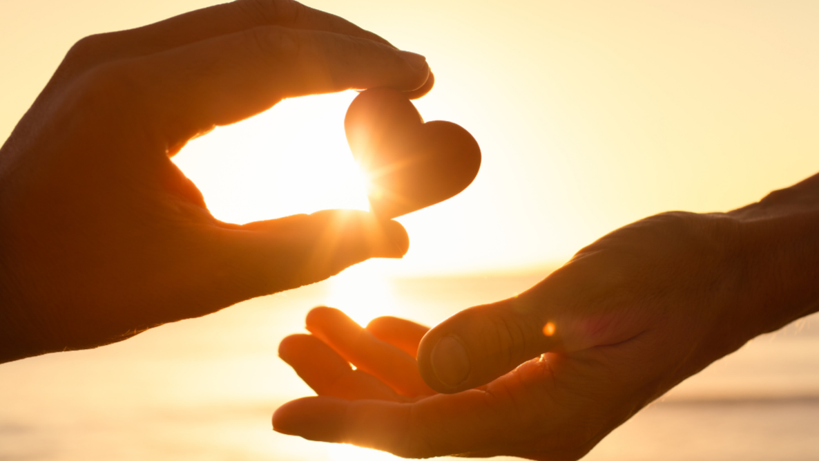 Silhouetted hands against a golden sunset, one gently passing a small heart-shaped object into the open palm of another, with sunlight glowing between them.