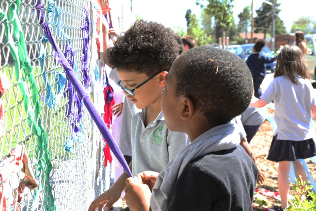 Students were shown how to create a fence "drawing" with fabric, and were supported to make their own creations. Divided into teams, these elementary school students invented stories, images, and words with multi-colored fabrics and a chain link fen