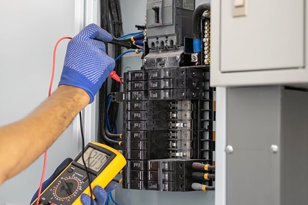 A person wearing blue gloves is using a digital multimeter to test electrical wires inside an electrical panel.