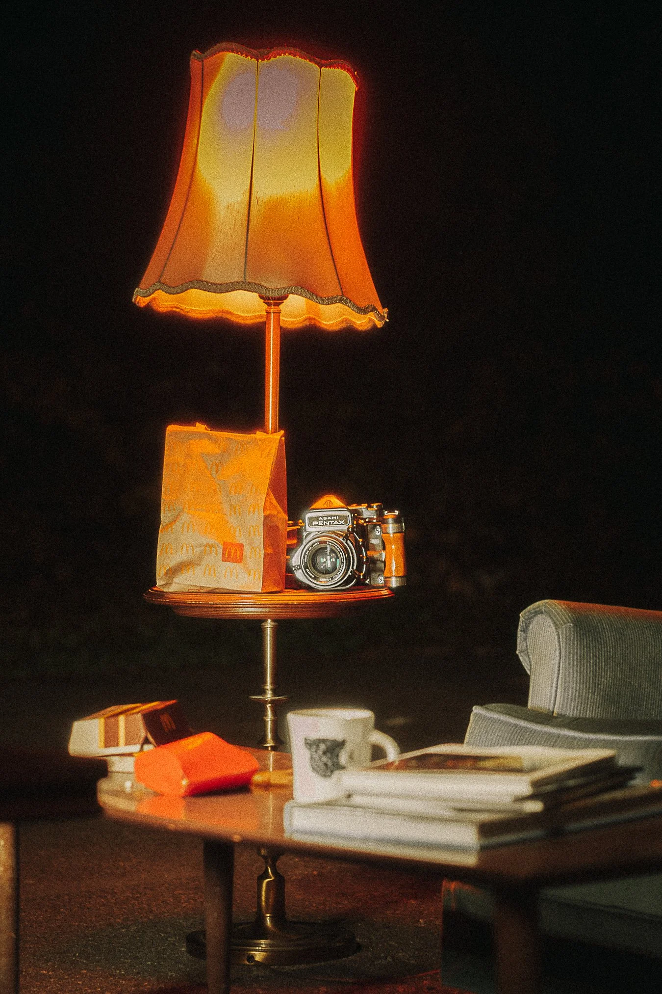 A table with various items, including a McDonald's paper bag, a camera, a coffee mug, and books, with a lamp made from an upside-down lampshade and a camera standing on a platform.
