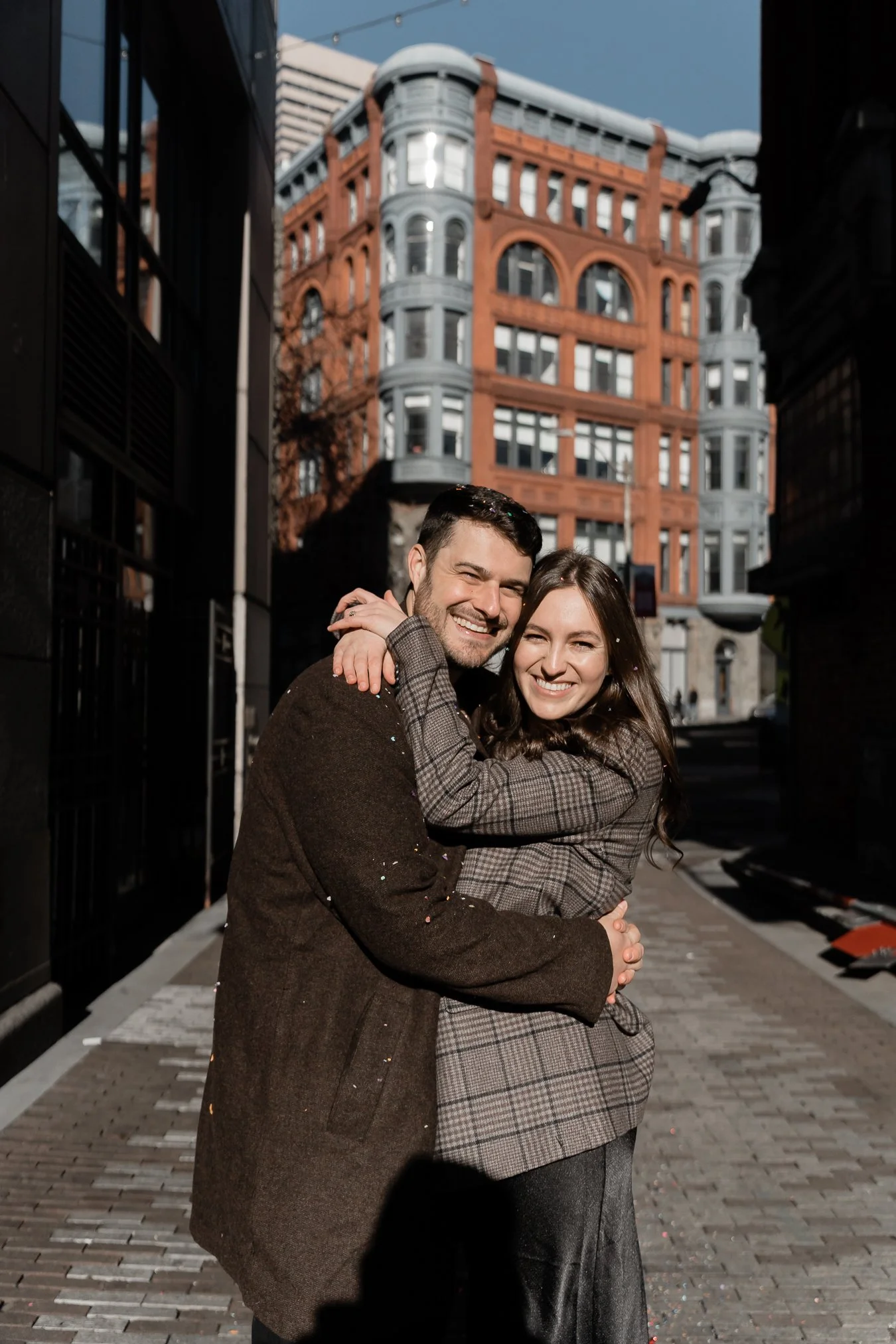 A smiling couple embracing each other outdoors in an urban area, with a historic red-brick building in the background.
