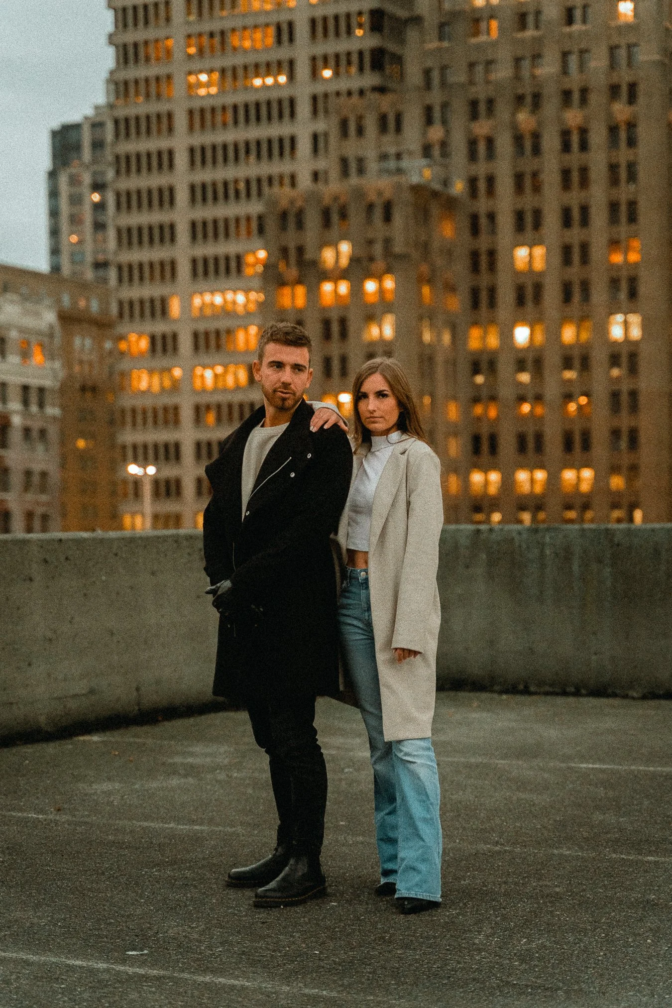 A young man and woman standing together on a rooftop with city skyscrapers in the background during dusk. The man is wearing a black coat and the woman is in a long cream coat and jeans.