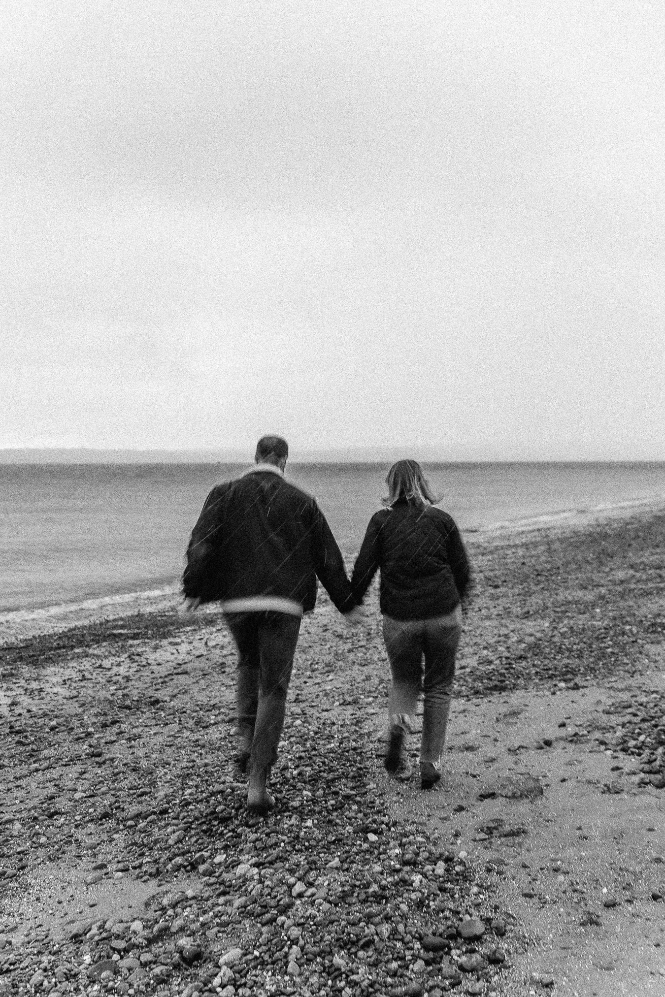 A black and white photo of a couple walking hand in hand on a pebble beach near the ocean.