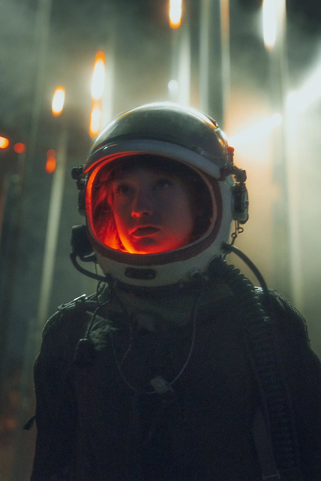 A woman wearing a space helmet looks upwards in a dimly lit environment with warm lights and a foggy atmosphere.